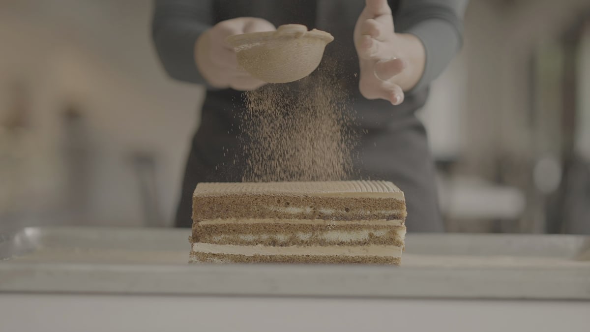 Person dusting powdered sugar onto a layered cake on a white surface, ungraded original footage before the CW CONTRAST LUT