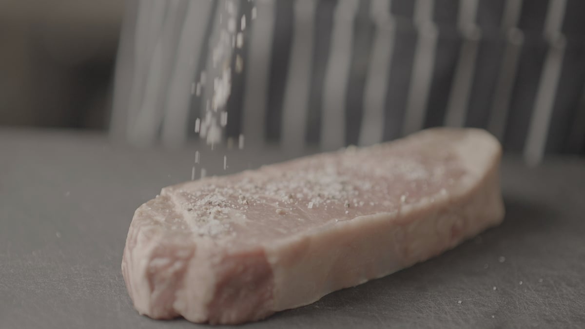 Raw pork tenderloin being seasoned with salt from a shaker on a gray surface, ungraded original footage before the CW COOL LUT
