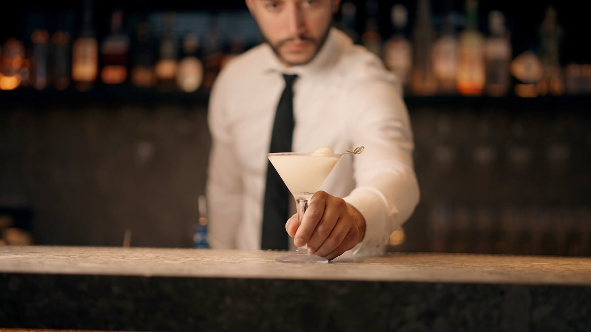 Bartender in white shirt and tie serving a cocktail at an upscale bar with ambient lighting, graded with the CW FOOD + PEOPLE LUT