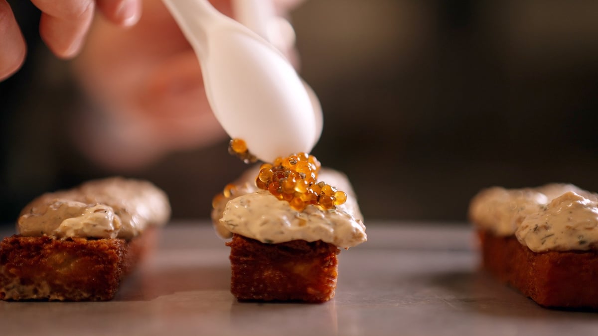 Hand piping gold-colored frosting onto a red velvet cupcake with other decorated cupcakes beside it, graded with the CW MAIN LUT