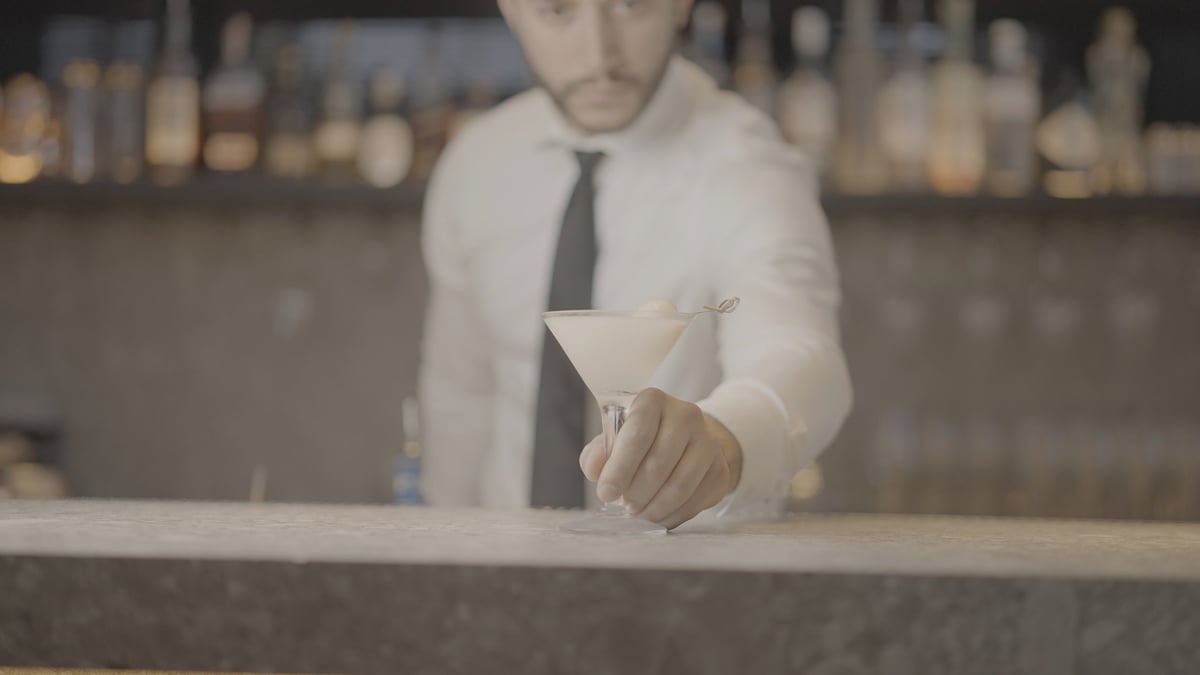 Bartender in white shirt and tie pouring a drink behind a bar counter with blurred bottles in background, ungraded original footage before the CW FOOD + PEOPLE LUT