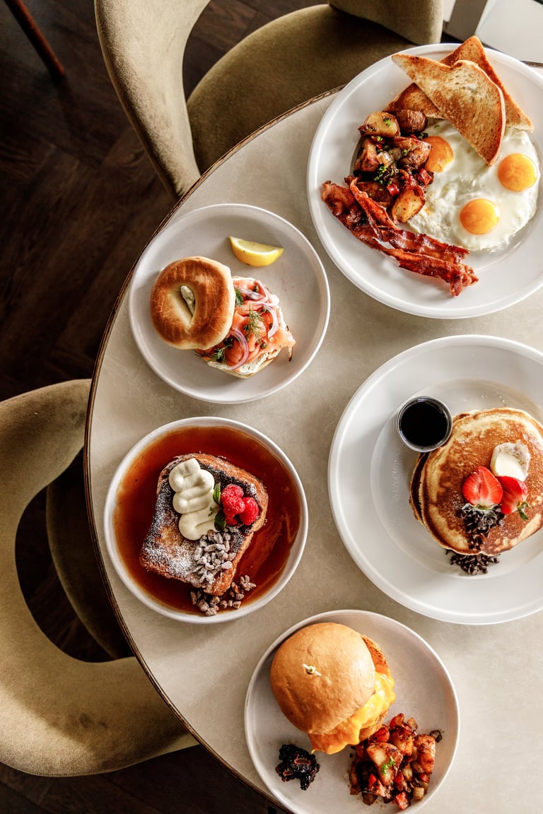 Overhead view of five white plates with various breakfast dishes including pancakes, eggs, bacon, and pastries on a light table