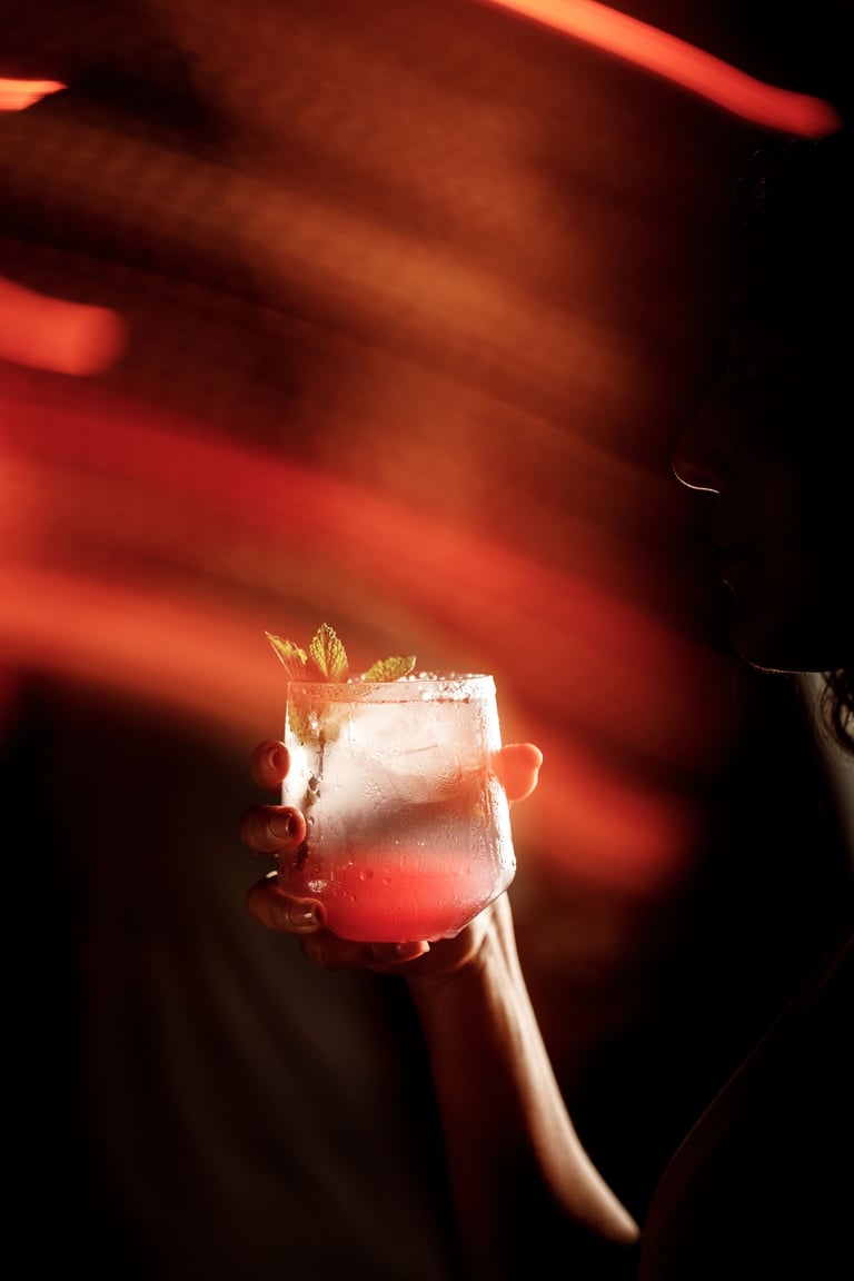 Hand holding a glass of icy pink cocktail with mint garnish against a warm orange and red bokeh background