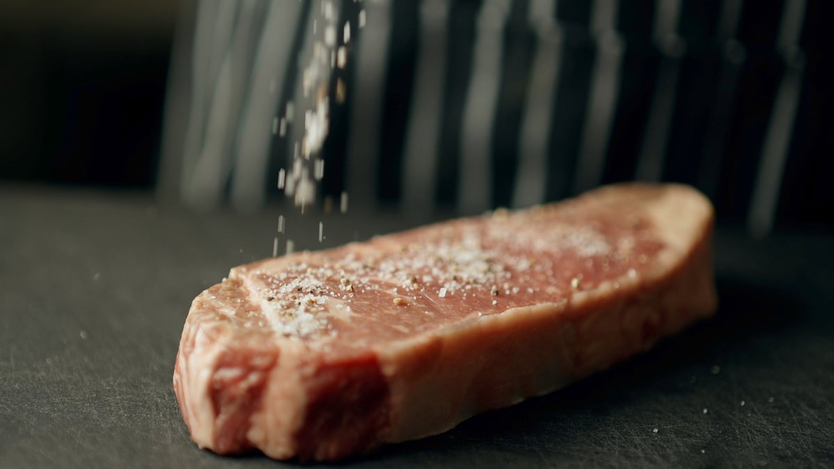 Salt being sprinkled onto a raw steak on a dark cooking surface, graded with the CW COOL LUT