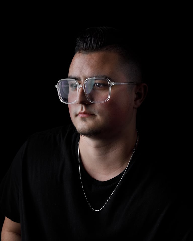 Man wearing clear-framed glasses and a black t-shirt with a silver chain necklace against a dark background