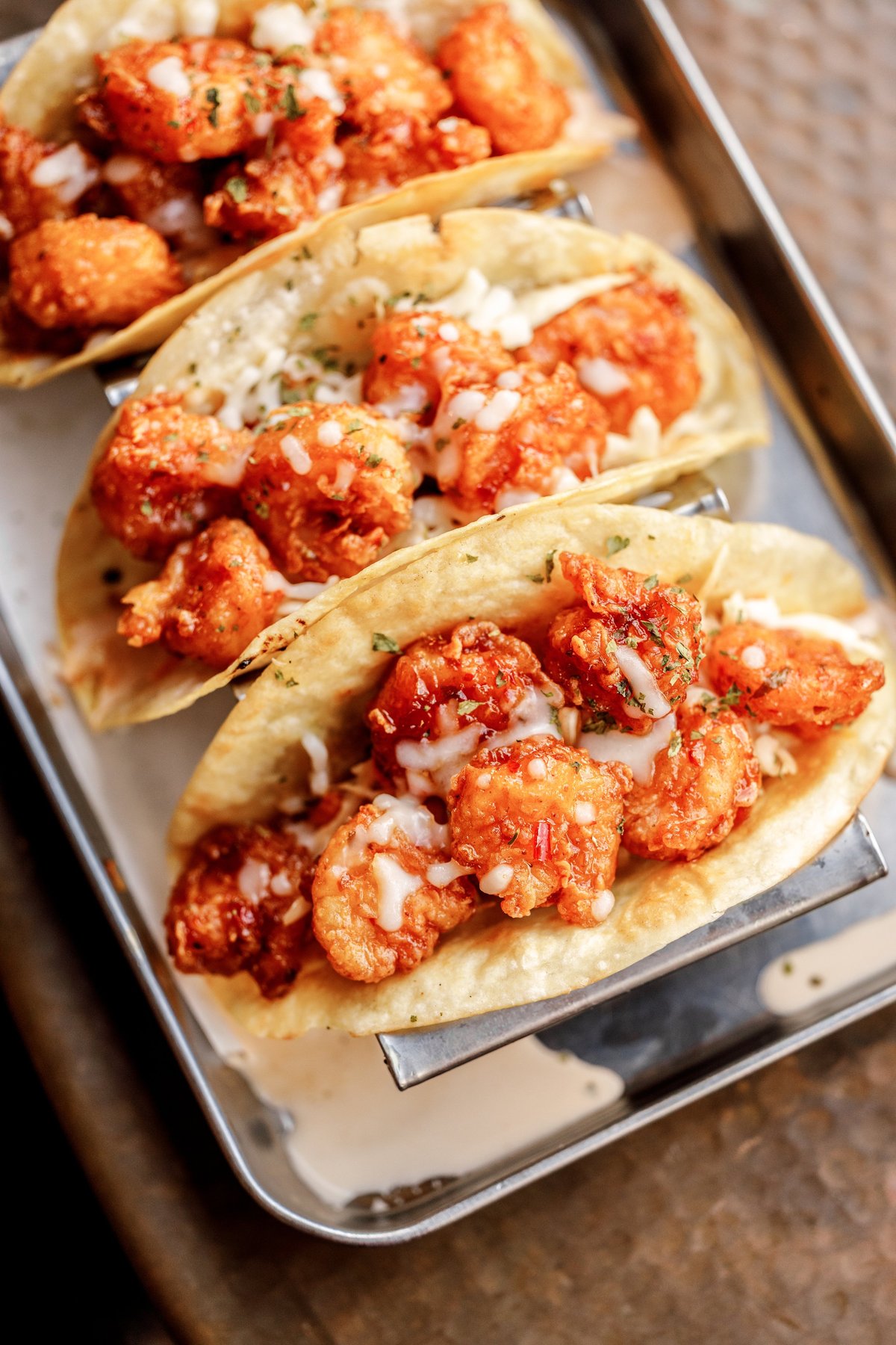 Three crispy shrimp tacos with spiced red coating, white sauce, and diced onions on soft flour tortillas served on a metal platter