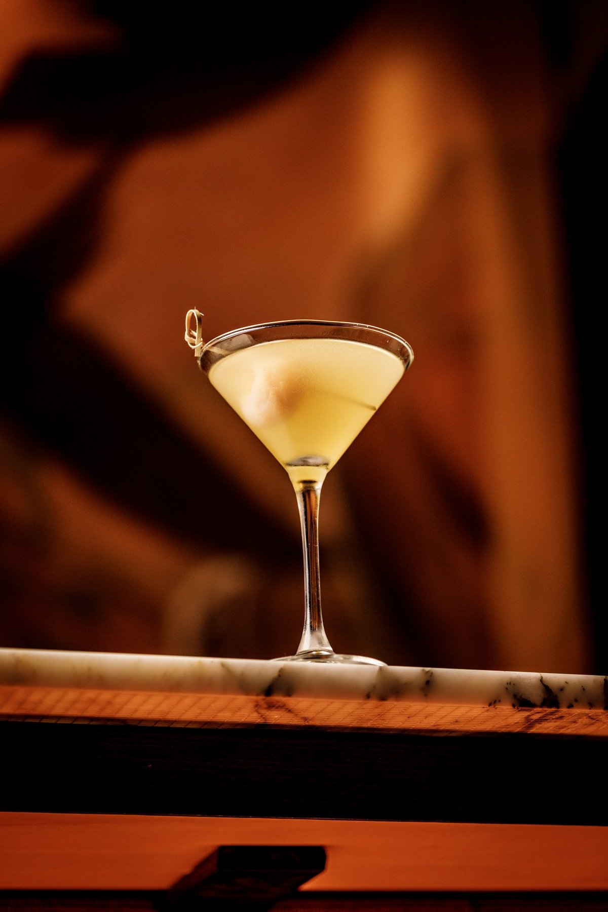 A pale yellow cocktail in a martini glass with a cocktail pick garnish, set against a warm amber-toned blurred background.